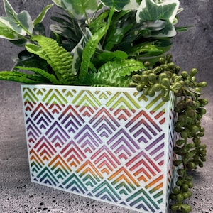 Puede incluir: Una maceta rectangular blanca con un colorido patrón geométrico en tonos de rojo, naranja, amarillo, verde, morado y azul. La maceta está llena de plantas de hojas verdes y blancas y bayas verdes.