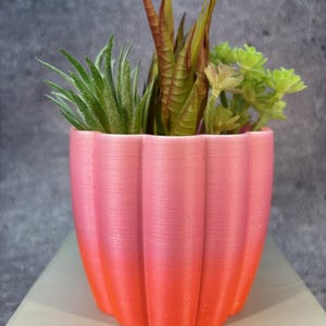 May include: A pink and orange ombre planter with a ribbed design, filled with a variety of green and brown artificial succulents.