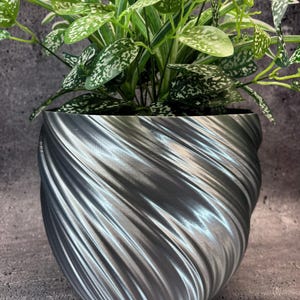 Peut inclure: Un pot de fleurs décoratif argenté avec un motif tourbillonnant, contenant une plante verte vibrante avec des feuilles tachetées de blanc. Le pot est en matériau métallique et repose sur une surface grise. La plante ajoute une touche naturelle.