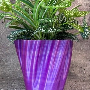 May include: A purple iridescent planter with a wavy design, holding a green plant with white speckles on the leaves.