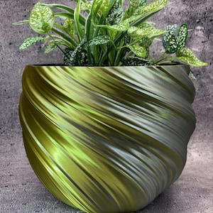 Puede incluir: Un macetero de espiral verde y plateado con una planta verde en su interior. El macetero está hecho de un material liso y brillante y tiene un diseño único y moderno.