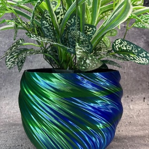 May include: A vibrant, two-toned planter with a swirling design, featuring shades of green and blue. The planter holds a lush houseplant with green and white speckled leaves. The pot's glossy finish adds a modern touch to the decor.