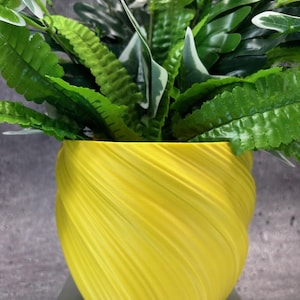 May include: A yellow ceramic planter with a swirling, textured design. The planter is filled with green leafy plants.