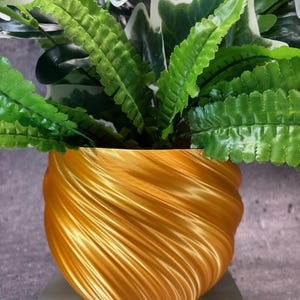 Puede incluir: Una maceta de cerámica dorada con un patrón de remolino y una planta verde con hojas.