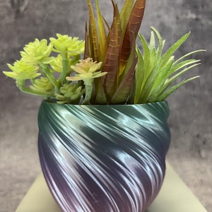 Op de afbeelding: Een kleine, ronde plantenpot met een spiraalvormige, geribbelde textuur met een verloop van metallic groene, paarse en zilveren tinten. De pot bevat een arrangement van kunstmatige vetplanten in tinten lichtgroen, geelgroen en roodbruin.