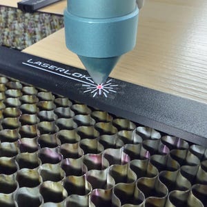 K&ouml;nnte beinhalten: Nahaufnahme einer Lasergravurmaschine in Betrieb. Die D&uuml;se der Maschine ist auf eine Holzoberfl&auml;che gerichtet und erzeugt ein Design. Der Text "LASERLOK" ist auf einer schwarzen F&uuml;hrungsschiene zusammen mit mm-Ma&szlig;en zu sehen.