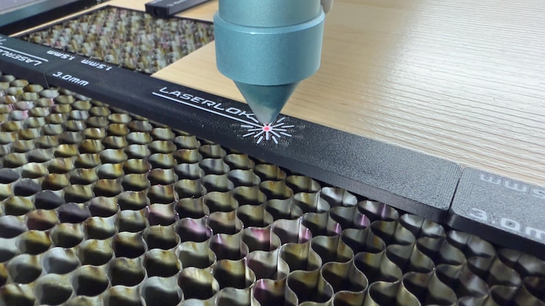 K&ouml;nnte beinhalten: Nahaufnahme einer Lasergravurmaschine in Betrieb. Die D&uuml;se der Maschine ist auf eine Holzoberfl&auml;che gerichtet und erzeugt ein Design. Der Text "LASERLOK" ist auf einer schwarzen F&uuml;hrungsschiene zusammen mit mm-Ma&szlig;en zu sehen.