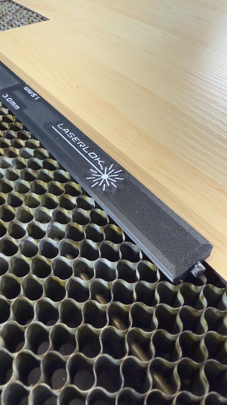 K&ouml;nnte beinhalten: Ein schwarzes Laserlok-Lineal mit wei&szlig;em Text und Sternlogo, misst 1,5 mm und 3,0 mm, liegt auf einer hellen Holzoberfl&auml;che und einem wabenf&ouml;rmigen Metallgitter. Das Lineal ist &uuml;ber das Holz geneigt.