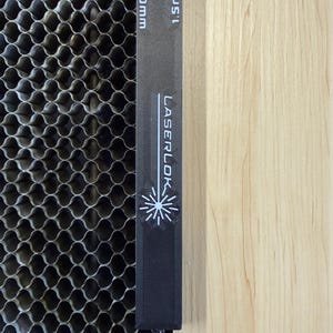 K&ouml;nnte beinhalten: Ein schwarzes Lineal mit dem Text "LASERLOK" und einem Schneeflocken-Design, mit den Ma&szlig;en 3,0 mm und 1,5 mm. Das Lineal befindet sich neben einem metallischen Wabenmuster und einer hellen Holzoberfl&auml;che.
