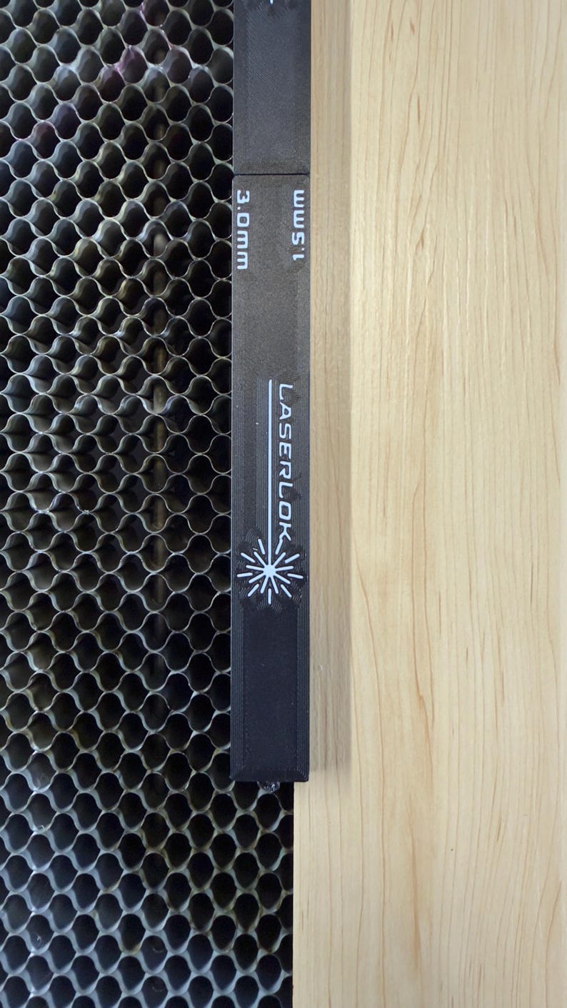 K&ouml;nnte beinhalten: Ein schwarzes Lineal mit dem Text "LASERLOK" und einem Schneeflocken-Design, mit den Ma&szlig;en 3,0 mm und 1,5 mm. Das Lineal befindet sich neben einem metallischen Wabenmuster und einer hellen Holzoberfl&auml;che.