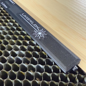 Puede incluir: Una regla LaserLok negra con texto blanco y un logotipo de estrella descansa sobre una superficie de madera clara y una rejilla metálica de panal. La regla tiene medidas de 1,5 mm y 3,0 mm.