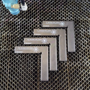May include: Four grey L-shaped metal Laserlok tools, each with a white logo and the text "1.5mm". The tools are arranged on a honeycomb metal surface. A blue and silver machine component is visible in the upper left corner.