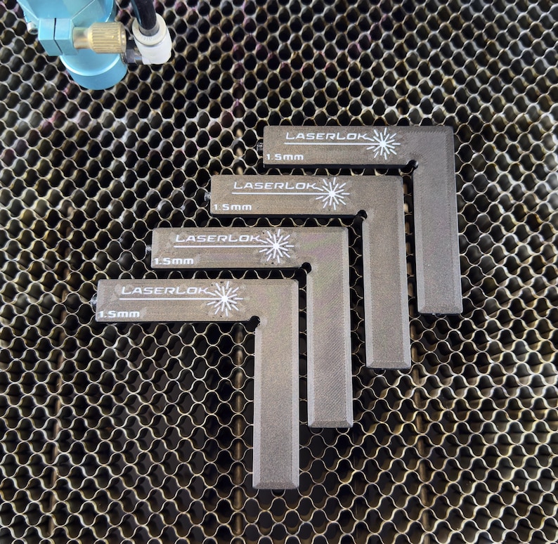 May include: Four grey L-shaped metal Laserlok tools, each with a white logo and the text "1.5mm". The tools are arranged on a honeycomb metal surface. A blue and silver machine component is visible in the upper left corner.