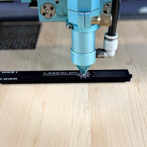 K&ouml;nnte beinhalten: Nahaufnahme einer Lasergravurmaschine in Betrieb. Der blaue Kopf der Maschine ist auf ein schwarzes Lineal mit dem Text "LASERLOK" und Ma&szlig;en fokussiert. Der Laser schneidet ein Design in das Lineal, das auf einer hellen Holzoberfl&auml;che liegt.
