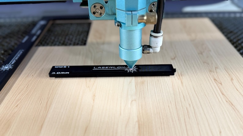 K&ouml;nnte beinhalten: Nahaufnahme einer Lasergravurmaschine in Betrieb. Der blaue Kopf der Maschine ist auf ein schwarzes Lineal mit dem Text "LASERLOK" und Ma&szlig;en fokussiert. Der Laser schneidet ein Design in das Lineal, das auf einer hellen Holzoberfl&auml;che liegt.