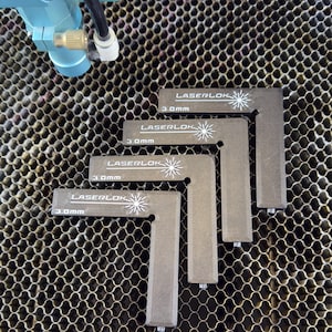 May include: Four grey, L-shaped LaserLok corner pieces, each with the text "LASERLOK" and "3.0mm" printed in white. The pieces are arranged on a honeycomb metal surface, with a blue machine component visible in the upper left corner.