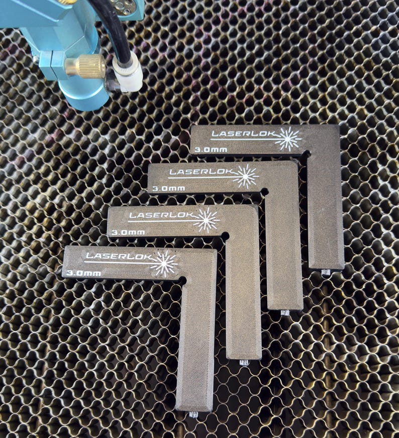 May include: Four grey, L-shaped LaserLok corner pieces, each with the text "LASERLOK" and "3.0mm" printed in white. The pieces are arranged on a honeycomb metal surface, with a blue machine component visible in the upper left corner.