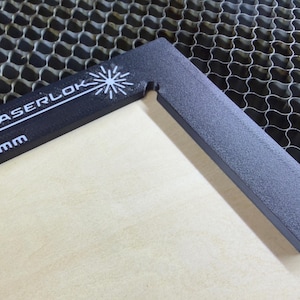 Puede incluir: Una escuadra de esquina negra Laserlok de 3,0 mm descansa sobre una superficie de madera de color claro. La herramienta tiene el texto blanco "LASERLOK" y "3,0 mm". La herramienta está colocada en un ángulo de 90 grados.