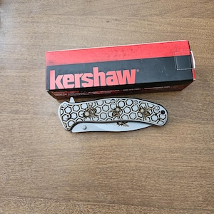 Puede incluir: Una navaja plegable plateada con un dise&ntilde;o de abeja y la marca "Kershaw" en la caja. La navaja presenta un patr&oacute;n de panal y ilustraciones de abejas. La caja es roja y negra.