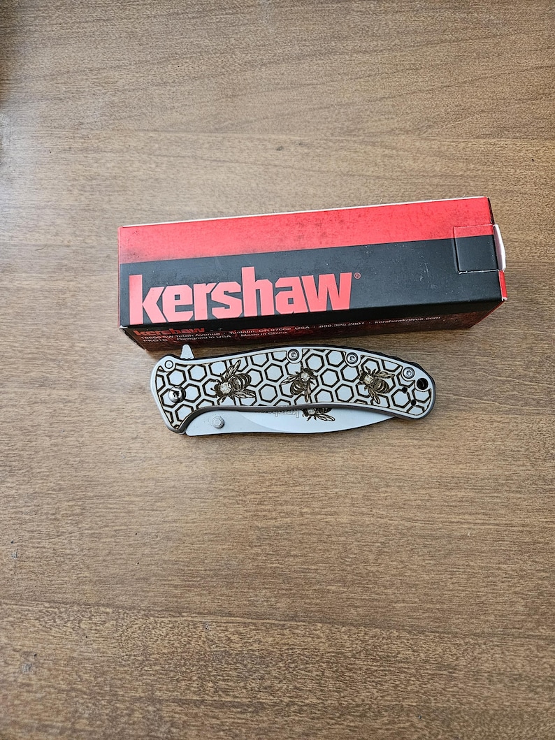 Puede incluir: Una navaja plegable plateada con un dise&ntilde;o de abeja y la marca "Kershaw" en la caja. La navaja presenta un patr&oacute;n de panal y ilustraciones de abejas. La caja es roja y negra.