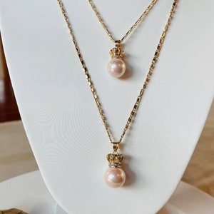 May include: A gold necklace with two pendants featuring a pearl and a crown. The necklace is paired with matching earrings with pearl and crown accents.