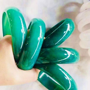 May include: A set of five green jade bangles, stacked and held in a hand. The bangles are smooth and polished, with a glossy finish.