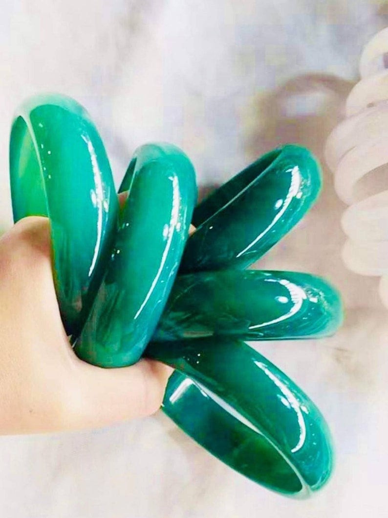 May include: A set of five green jade bangles, stacked and held in a hand. The bangles are smooth and polished, with a glossy finish.