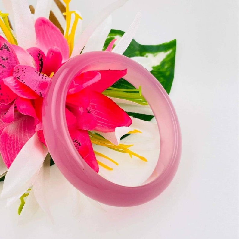 May include: A pink bangle bracelet made of smooth, polished stone. The bracelet is displayed on a white background with pink flowers.