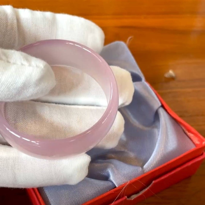 May include: A pale pink jade bangle bracelet in a blue and red box. The bangle is smooth and polished.