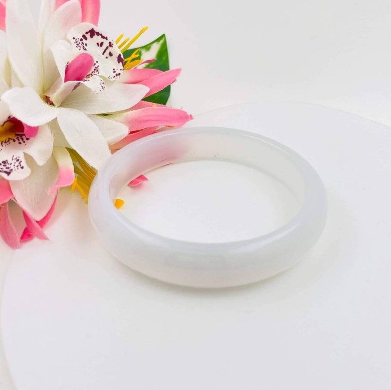 May include: A white jade bangle bracelet. The bracelet is smooth and polished, with a slightly rounded shape. It is a simple and elegant piece of jewelry.