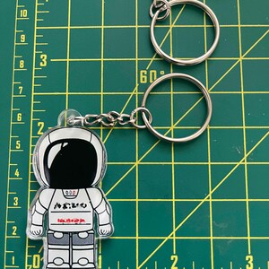 May include: A clear acrylic astronaut keychain with a silver chain and keyring. The astronaut is white with black accents and the word "NASA" in red. The keychain is approximately 3 inches tall.