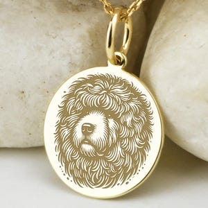 May include: Gold pendant necklace with a detailed dog face engraving. The circular pendant hangs from a fine chain with small ball details. The necklace is displayed on a white stone surface.