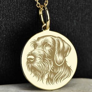 Könnte beinhalten: Eine goldfarbene Halskette mit Anhänger, die ein detailliertes, eingraviertes Porträt eines Hundekopfes zeigt. Der runde Anhänger hängt an einer zarten Goldkette mit kleinen, runden Perlen. Der Hintergrund ist dunkel.