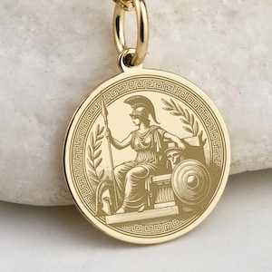 May include: A gold-colored pendant necklace featuring a circular charm with an engraved image of a seated figure in classical attire, holding a spear and shield. The charm is surrounded by a laurel wreath and a Greek key border. The chain has small ball accents.