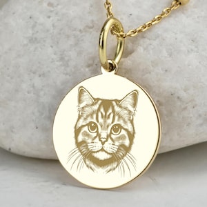 Może przedstawiać: Złoty naszyjnik z okrągłą zawieszką z wygrawerowaną twarzą kota. Zawieszka zawieszona jest na delikatnym łańcuszku z małymi, kulistymi koralikami. Cechy kota są szczegółowe, w tym wąsy i oczy.