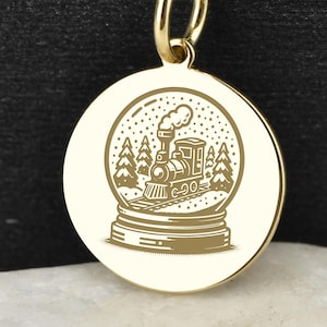 May include: A gold-colored pendant necklace featuring a round charm with a detailed engraving of a snow globe. Inside the snow globe is a train, trees, and falling snow. The necklace chain is also gold and has small ball accents.