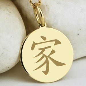 May include: Gold-colored pendant necklace with a circular charm featuring a Chinese character. The pendant hangs from a delicate chain with small, round beads. The necklace is displayed against a backdrop of white stones.