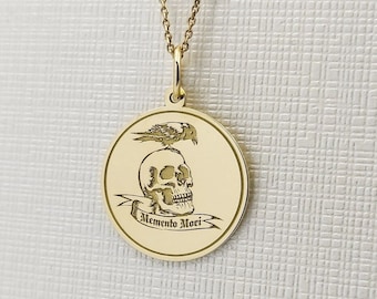 Könnte beinhalten: Eine goldene, kreisförmige Anhängerkette mit einer Gravur, die einen Totenkopf mit einem darauf sitzenden Vogel zeigt. Unter dem Totenkopf befindet sich ein Banner mit der Inschrift Memento Mori. Der Anhänger hängt an einer feinen Goldkette.
