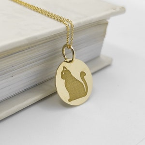 May include: A gold necklace with a round pendant featuring a silhouette of a cat. The pendant is hanging from a delicate chain.
