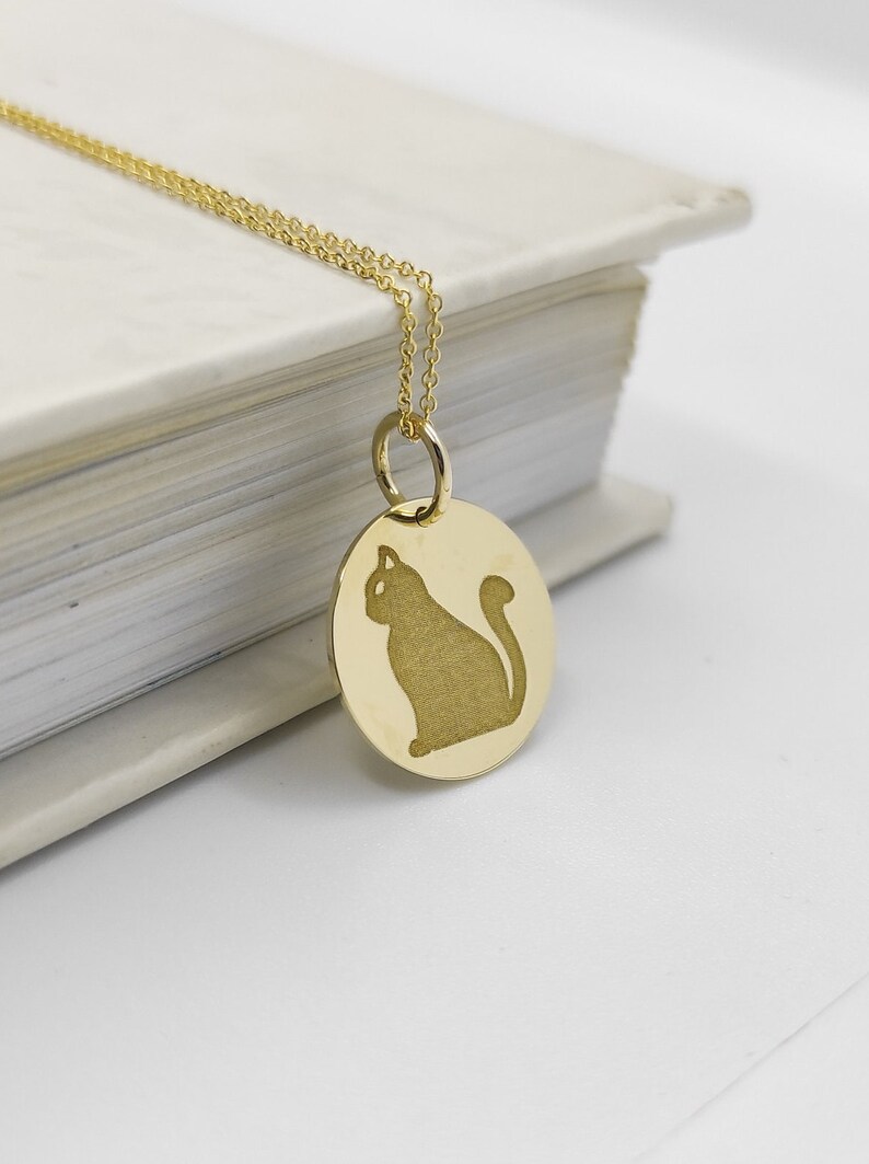 May include: A gold necklace with a round pendant featuring a silhouette of a cat. The pendant is hanging from a delicate chain.