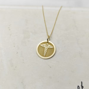 K&ouml;nnte beinhalten: Eine goldene Halskette mit einem runden Anh&auml;nger, auf dem ein medizinisches Symbol, der Caduceus, in Wei&szlig; graviert ist.