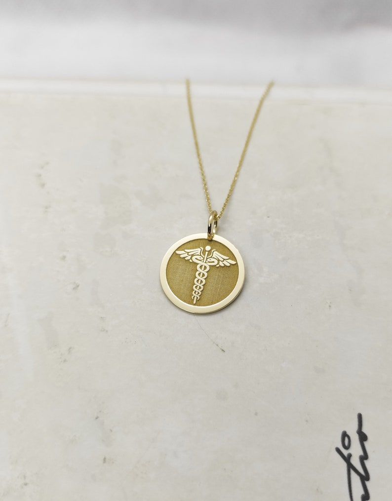 K&ouml;nnte beinhalten: Eine goldene Halskette mit einem runden Anh&auml;nger, auf dem ein medizinisches Symbol, der Caduceus, in Wei&szlig; graviert ist.