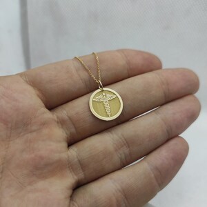 K&ouml;nnte beinhalten: Eine goldene Halskette mit einem runden Anh&auml;nger, der ein medizinisches Symbol zeigt, den Caduceus, mit Fl&uuml;geln und einer Schlange, die um einen Stab gewickelt ist.