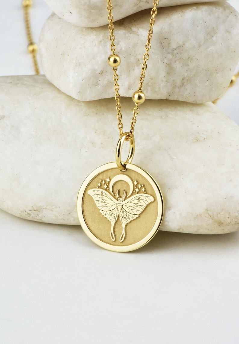 May include: Gold necklace with a round pendant featuring a detailed engraving of a moth with wings spread and a crescent moon above it.