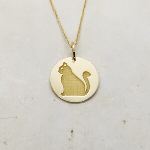 May include: A gold necklace with a round pendant featuring a silhouette of a cat. The pendant is attached to a delicate gold chain.