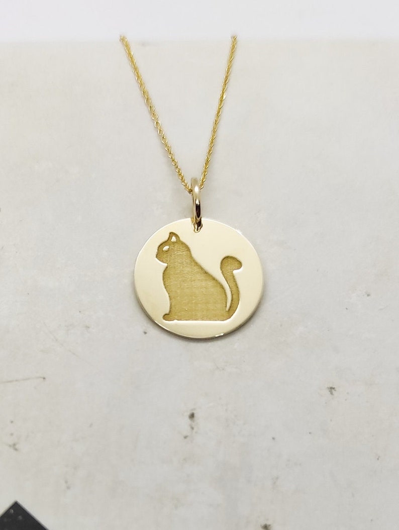 May include: A gold necklace with a round pendant featuring a silhouette of a cat. The pendant is attached to a delicate gold chain.