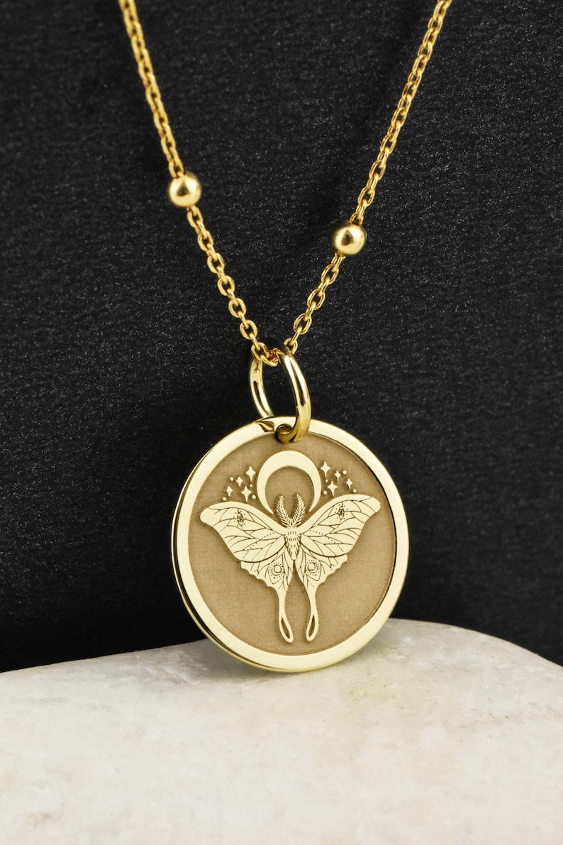 May include: Gold necklace with a round pendant featuring a detailed engraving of a moth with a crescent moon and stars.