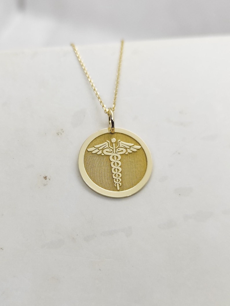 K&ouml;nnte beinhalten: Eine goldene Halskette mit einem runden Anh&auml;nger, der ein medizinisches Symbol, den Caduceus, mit einer Schlange und Fl&uuml;geln zeigt.