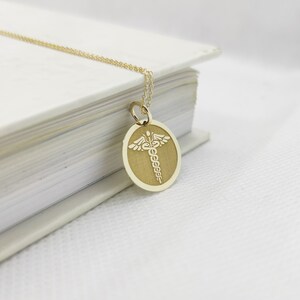 K&ouml;nnte beinhalten: Eine goldene Halskette mit einem runden Anh&auml;nger, auf dem ein medizinisches Symbol, der Caduceus, in Wei&szlig; graviert ist.