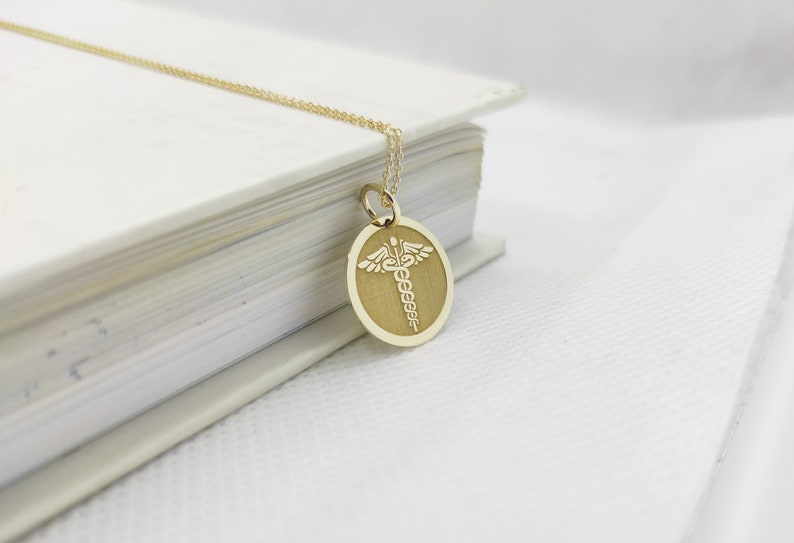 K&ouml;nnte beinhalten: Eine goldene Halskette mit einem runden Anh&auml;nger, auf dem ein medizinisches Symbol, der Caduceus, in Wei&szlig; graviert ist.